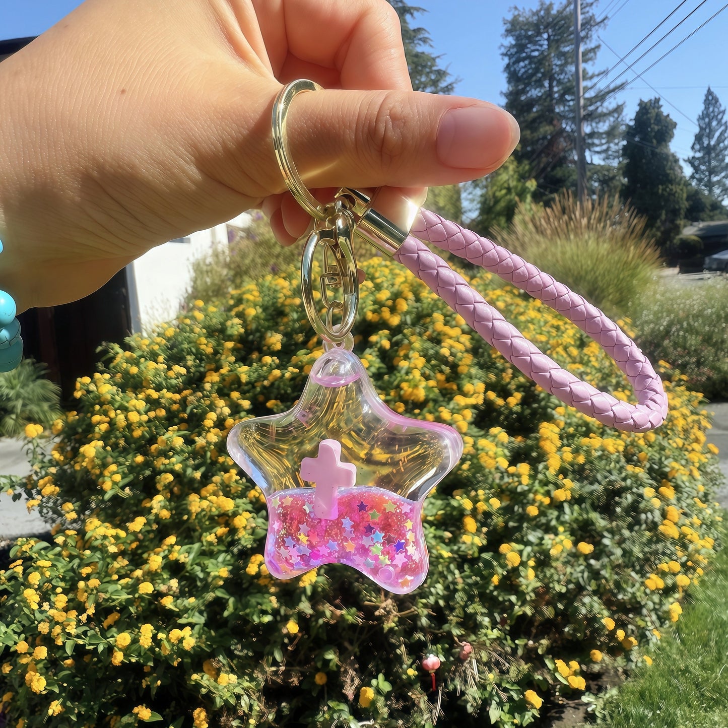 Hand holding a star-shaped keychain with a pink bow against a background of yellow flowers and greenery.